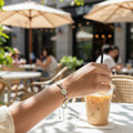 BlingbyAnvika silver daisy charm bracelet with white and yellow floral motifs worn at an outdoor cafe table.